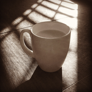 Photo of a white coffee mug sitting on a dark table with light shining across the table showing the shadows of an open blind from the window.