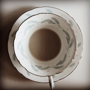 Photo looking down on the top of a china teacup with delicate leaves printed on the inside of the cup and around the edges of the saucer beneath the cup. The cup has a small amount of coffee with cream in the bottom.